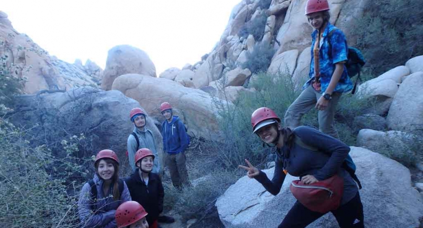 A group of people wearing helmets gather near large boulders. One of them gives the camera a peace sign. 